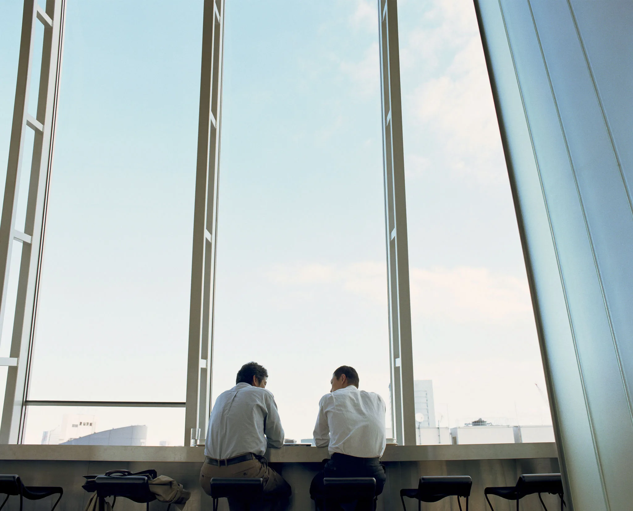 Two men in a high-rise office building sit in front of large open windows, overlooking the city below.